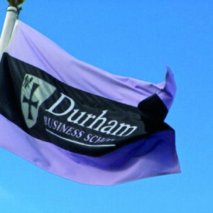 A custom flag flying with a blue sky behind. The flag is white with a black horizontal stripe with Durham Business School and the coat of arms of the School.
